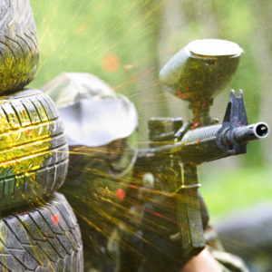 paintball sport player wearing protective mask aiming gun from shelter under gunfire attack with paint splash