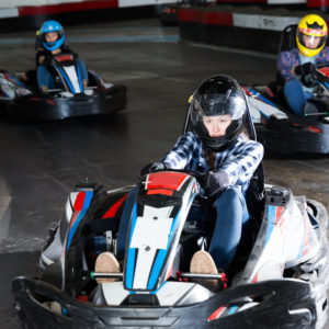 Group of male and females in helmets driving racing cars at kart track.