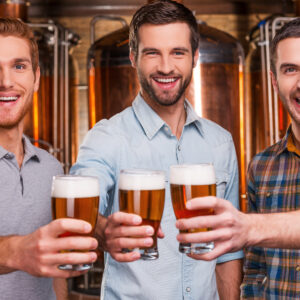 Cheers to you! Three cheerful young men in casual wear stretching out glasses with beer and smiling while standing in front of metal containers